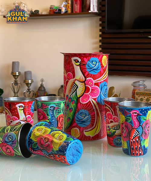 Colorful hand-painted metal tumblers and pitcher with floral and bird designs on a glass table
