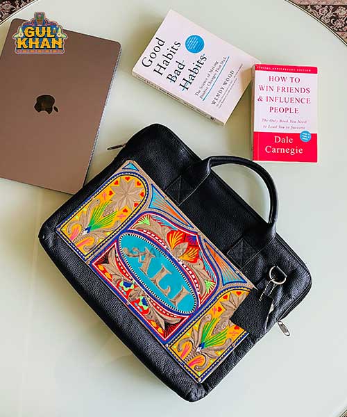 Black leather briefcase with colorful traditional embroidery labeled XL, next to an iPad and two self-help books on a white table