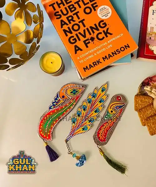 Colorful handmade bookmarks with intricate patterns placed near a yellow candle, golden decor, and an orange book by Mark Manson