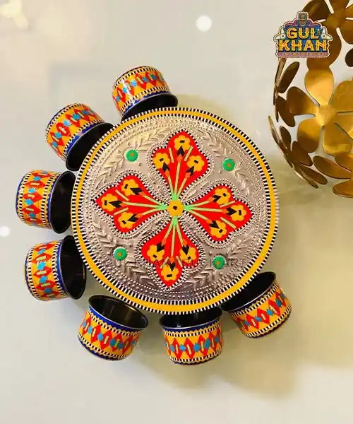 Traditional Gul Khan metal handi with engraved floral design and six colorful matching cups on a light surface